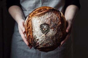 Mueller Communication's logo "branded" into the crust of a loaf of sourdough bread being held by a baker.